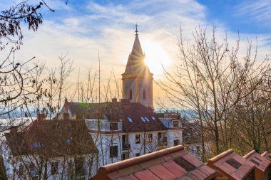 Çıplak kış ağaçları ve çatıların arasında gündoğumu manzaralı kilise kulesi.