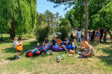 Bir grup insan parktaki şişme sandalyelerde oturuyor. Çocuklar oynuyor ve yetişkinler oturup konuşuyor. Sahne rahat ve eğlenceli. 1 Haziran 2024 Balti Moldova.
