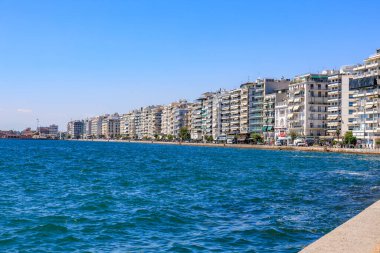 Mavi okyanusu ve binaları olan bir sahil. Binalar yüksek ve balkonları var. Sahil insanlarla dolu ve oturabilecekleri banklar var. 8 Ağustos 2024 Selanik Yunanistan