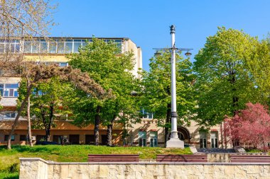 Modern binanın yakınında yemyeşil ağaçları ve bankları olan güneşli bir şehir parkı. 3 Mayıs 2025 Romanya.