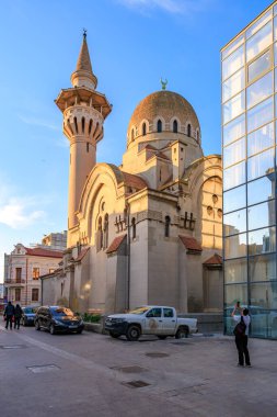Günbatımında şehir merkezinde minareli tarihi cami ve modern cam bina. 3 Mayıs 2025 Romanya.