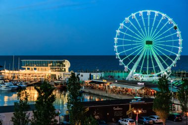 Limanda tekneleri olan deniz kenarındaki parlak dönme dolabın akşam manzarası. 3 Mayıs 2025 Romanya.