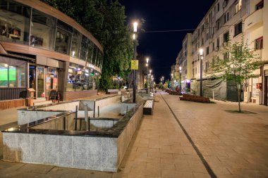 Geceleri dükkan ve banklarla aydınlatılmış şehir caddesi. 3 Mayıs 2025 Romanya.