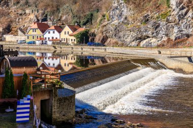 Nehir akan küçük bir kasaba. Nehir bir şelalenin üzerinden akıyor ve su kahverengi. Nehir boyunca park edilmiş birkaç ev ve birkaç araba var. 21 Mart 2025, Cesky Krumlov, Çek.