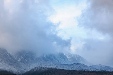 Bulutlu gökyüzünün altında karla kaplı tepeleri olan puslu dağ sıraları.