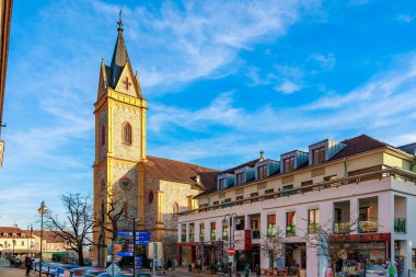 Mavi gökyüzü altında kilise ve modern binaları olan tarihi Avrupa meydanı. 21 Mart 2025, Hluboka Şatosu, Çek.