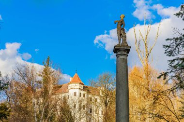 Klasik Avrupa mimarisi yakınlarında parlak mavi gökyüzüne karşı sütun üzerine tarihi heykel. 17 Mart 2025, Konopiste Şatosu, Çek Cumhuriyeti.