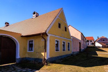 Rustic yellow cottage with red roofs and blue sky in quaint rural village setting. March 21, 2025, Holasovice, Czechia.