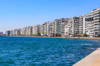 Mavi okyanusu ve binaları olan bir sahil. Binalar yüksek ve balkonları var. Sahil insanlarla dolu ve oturabilecekleri banklar var. 8 Ağustos 2024 Selanik Yunanistan