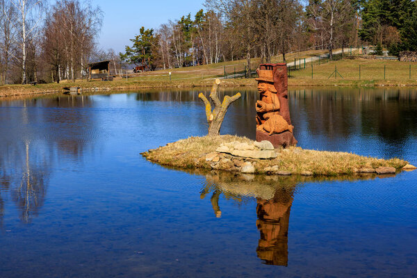 Wooden statue on island reflecting in tranquil lake with trees and blue sky. March 21, 2025, Holasovice, Czechia.