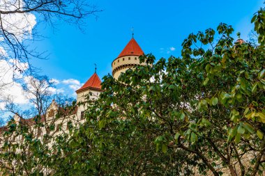 Historic stone castle with red turrets and leafy trees on a clear day.