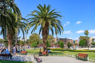 Sunny park with palm trees and people relaxing on benches in urban setting. August 12, 2025 Istanbul Turkey.