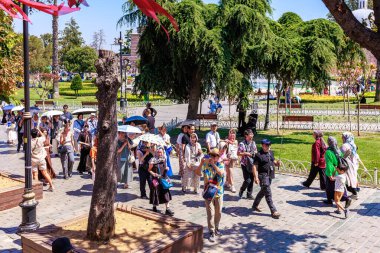 Diverse crowd walking in sunlit park with greenery and trees. August 12, 2025 Istanbul Turkey.