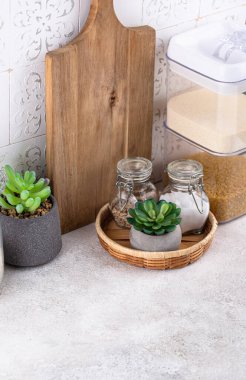 Mutfak tezgahının üstünde kavanozlar ve beyaz arka planda kesim tahtaları var.