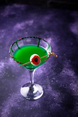 Vibrant green cocktail with red eyeball garnish against purple misty background clipart