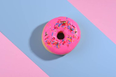 Parlak pembe bir donut renkli serpintilerle Geometrik pembe ve mavi bir arka planda durur.