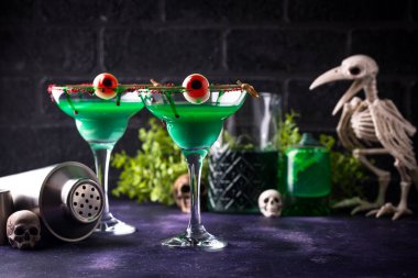 Cadılar Bayramı temalı yeşil kokteyller. Arka planda koyu renk göz süsleri ve iskelet süslemeleri var.