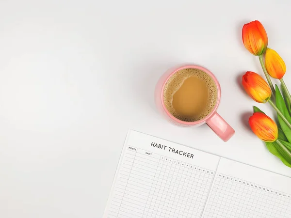 Top view or flat layout of Habit tracker book, cup of coffee and tulips on white background.