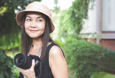 Asyalı bir kadının portresi, şapkasız ve siyah kolsuz, bahçede duruyor ve elinde video kamera, mutlu bir şekilde gülümsüyor..