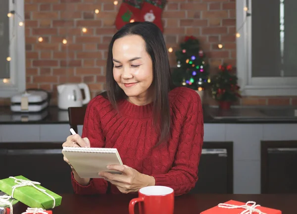 stock image Portrait of Asian woman wearing red knitted sweater sitting  at table with red cup of coffee and gift boxes  in the kitchen with Christmas decoration, using pen writing name list on notebook.