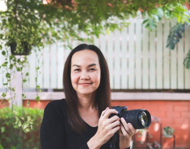 Asyalı bir kadının portresi, uzun kollu bluz giyiyor, bahçede dikiliyor, elinde video kamera, mutlu bir şekilde gülümsüyor..