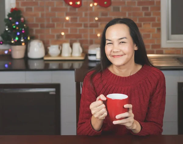 Portrait of Asian woman wearing red knitted sweater sitting in the kitchen with Christmas decoration, holding red cup of coffee, smiling and looking at camera.