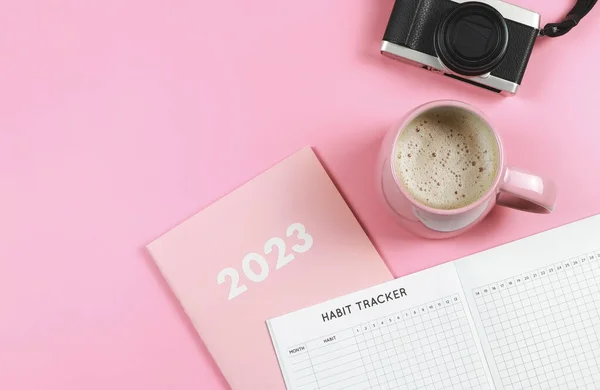 Top view or flat lay of habit tracker book on pink diary or planner 2023, pink cup of coffee and digital camera on pink background with copy space.