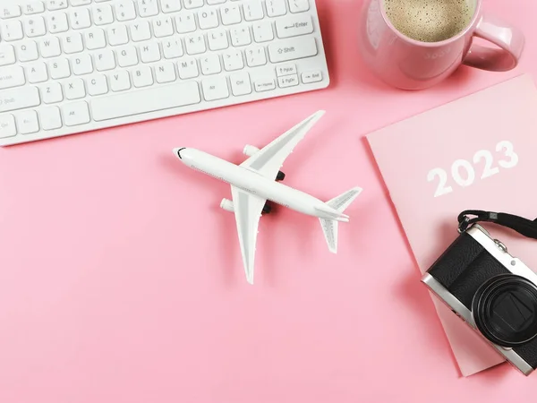 Top view or flat lay of computer keyboard, pink cup of coffee, airplane model, 2023 diary or planner and camera on  pink background with copy space. Business and Traveling concept.
