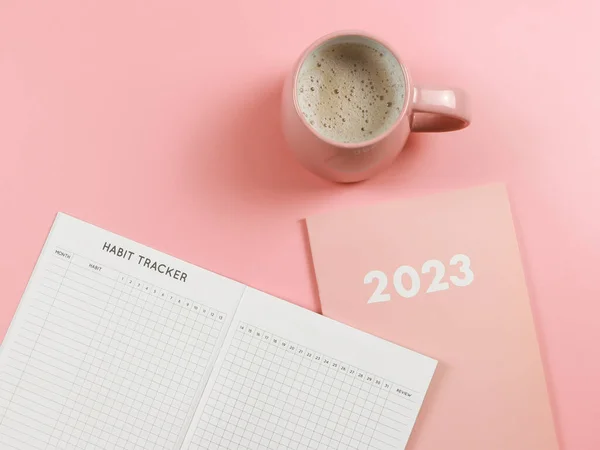 Top view or flat lay of habit tracker book on pink diary or planner 2023 and pink cup of coffee on pink background with copy space.