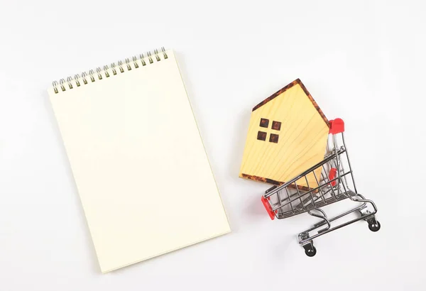 Top view or flat layout of wooden house model in shopping trolley or shopping cart with blank page opened notebook on white background, home purchase concept.