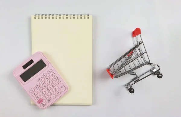 Top view or flat lay of pink calculator on blank page opened notebook and trolley or shopping cart on white background.. shopping budget, business and education concept.
