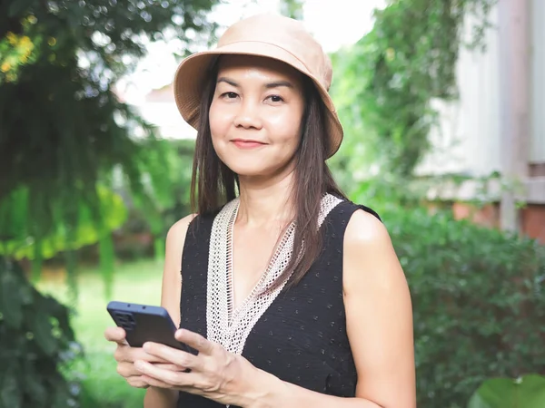 亚洲女人的画像 头戴帽子 头戴黑色无袖上衣 站在花园里拿着手机 看着相机 — 图库照片