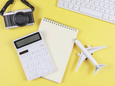 Sarı arkaplan, iş dünyası ve seyahat konsepti üzerine kopya alanı olan uçak modeli, bilgisayar klavyesi, not defteri, hesap makinesi ve dijital kameranın üst görünümü.
