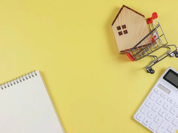 Top view or flat layout of wooden house model in shopping trolley, white calculator, blank page opened  notebook  on yellow  background with copy space, home purchase concept.