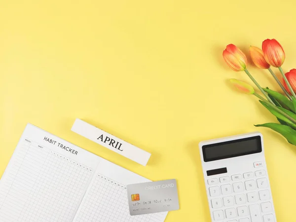 Top view or flat layout of Habit tracker book,  white calculator, credit card, wooden calendar April and tulips on yellow background.