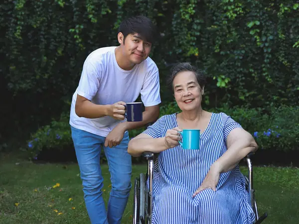 Portrait of Asian senior woman sitting on wheelchair, drinking coffee or tea with her son in the garden. looking at camera, smiling happily.