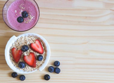 Kahvaltının üstü ya da üstü beyaz kasede bir bardak yaban mersinli smoothie ve yulaf ya da granola, taze yaban mersini, ahşap masada çilek. Sağlıklı kahvaltı kavramı.