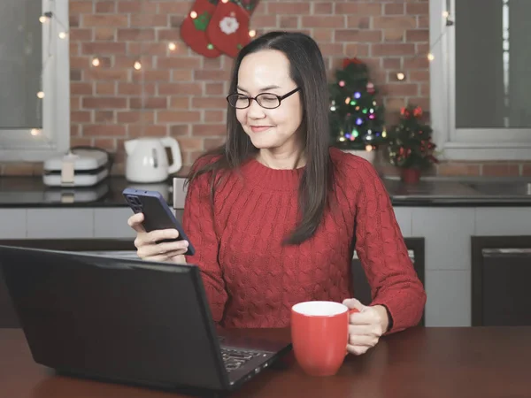 Portrait of Asian woman wearing eyeglasses ,red knitted sweater sitting with computer laptop  in ithe kitchen with Christmas decoration, holding  red cup of coffee , looking at mobile phone and smiling.