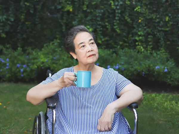 Portrait of Asian senior woman sitting on wheelchair, holding blue cup of tea or coffee in the garden.