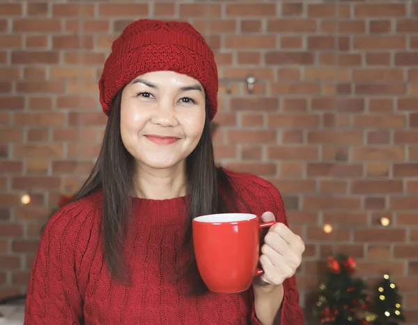 Portrait of Asian woman wearing red knitted sweater and hat standing in the kitchen decorated with Christmas tree drinking coffee from red mug, smiling happily and looking at camera.