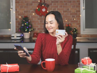 Kırmızı süveter giyen Asyalı bir kadının portresi mutfakta kırmızı kahve fincanı ve hediye kutusuyla oturuyor, Noel süslemesi, cep telefonu ve kredi kartı tutuyor, gülümsüyor ve kameraya bakıyor..