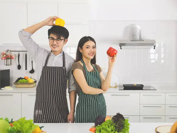 Young Asian couple preparing food in modern kitchen holding yellow and red capsicum smiling and look at the camera.