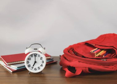 Kırmızılı okul çantasının önü, kırtasiye malzemeleri, kitaplar ve beyaz alarmlı saat. Saat 7 yönünde, ahşap masa üzerinde, beyaz duvar..