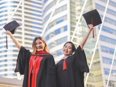 Mezuniyet elbisesi giymiş, elinde mezuniyet şapkası olan ve kameraya gülümseyen iki mutlu kadın. Şehir binası geçmişi. Eğitim, başarı ve arkadaşlık kavramı.