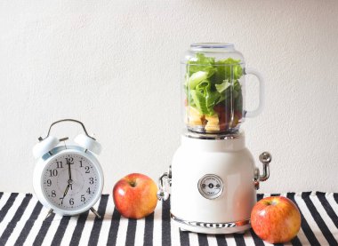 Beyaz vintage blender ya da meyve ve sebzeli smoothie makinesinin ön görüntüsü, beyaz vintage alarm saati saat 7 'yi, siyah beyaz çizgili masa örtüsü ve beyaz duvar örtüsü üzerinde 2 elmayı gösteriyor.