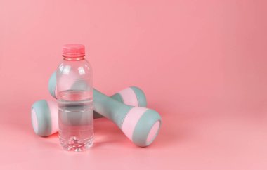 Pembe su şişesinin önü ve pembe arka planda izole edilmiş iki dambıl. Sağlık ve zindelik kavramı.