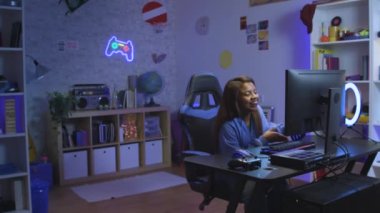 Mutlu genç Latin kız okuldan eve geliyor ve bilgisayar başında oturmuş neon ışıkları açık halde odasında video oyunları oynuyor. Etkileyici blog. Kırmızı kamerayla çekilmiş. Yüksek kalite 4k