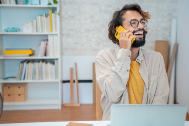 Genç yaratıcı girişimci telefonda konuşuyor. Evdeki modern çalışma alanında gülerek gülüyor. Serbest çalışan adam beyaz ve sarı renkli ofis ve fotokopi alanı olan müşteriyle konuşmaktan mutlu. Yüksek