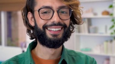 Modern işyerinde kameraya gülümseyen genç yaratıcı girişimcinin portresi. Ev ofisinde gözlüklü ve yeşil ceketli serbest çalışan bir adam. Yüksek kaliteli 4K video görüntüleri