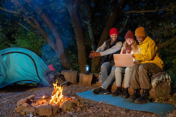 Happy multiethnic friends having fun playing music and enjoying bonfire camping in nature at night. Group of people chilling at fire in the evening, camping in the forest near lake. High quality photo